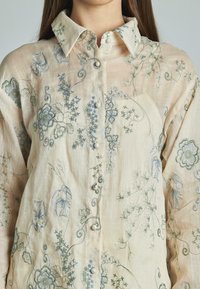 Camicia leggera in lino con ricami floreali in blu e verde. Presenta un colletto e bottoni simili a perle lungo la parte anteriore.