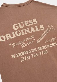 Bruine katoenen T-shirt met crème-kleurige print. De tekst bevat "GUESS ORIGINALS," "Professional Rookie," en "Hardware Services" met een schroefafbeelding.