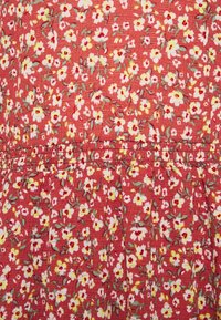 Tissu rouge à motifs floraux avec de petites fleurs blanches et jaunes sur un fond texturé, avec des plis et des détails de couture froncée.