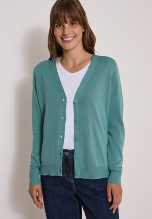 Kvinde smilende iført en teal knap-cardigan over en hvid skjorte og mørkeblå jeans, stående mod en ensfarvet lys baggrund.