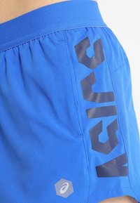 Blå sportshorts med elastisk midja, med en stor svart logotyp och grafik på sidan, slät tygtextur.