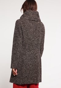 Brun tweedfrakke med høj krave, lange ærmer og et struktureret mønster. Stoffet har et spraglet udseende og en løs pasform.
