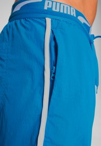 Pantalones cortos azules hechos de un tejido ligero y texturizado, con un bolsillo con cremallera blanca y una cinturilla elástica con la marca "PUMA".