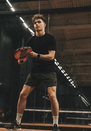 Young man in black shirt and shorts holding a paddle, preparing to play paddle tennis on indoor court with glass walls.