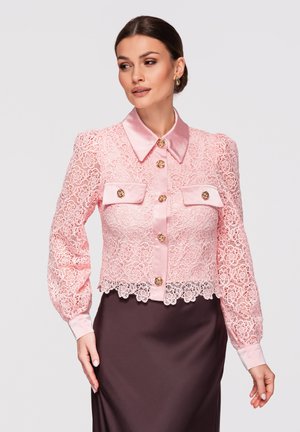 Kvinde iført en lyserød blondebluse med knapper, satin krave og manchetter, kombineret med et mørkebrun skørt, poserer mod en ensfarvet baggrund.