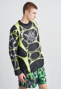 Μαύρο και νεον πράσινο μακρυμάνικο τοπ με αφηρημένο σχέδιο και λογότυπο, σε συνδυασμό με μαύρο σορτς που έχει πράσινο, υφασμάτινο μοτίβο.