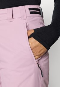 Pantaloni exteriori roz deschis, cu buzunar cu fermoar, talie ajustabilă și accente negre. Textură netedă, croială adaptată pentru funcționalitate.