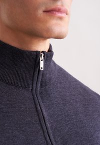 Primo piano della parte inferiore del viso e del collo di un uomo che indossa un maglione con zip di un blu navy scuro, con un colletto a coste.