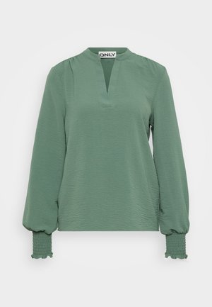 Blouse verte à manches longues avec un décolleté en V, en tissu texturé, et des manches bouffantes ornées de smocks au niveau des poignets.