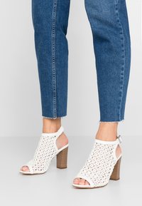 Bílé otevřené sandály na podpatku vyrobené z perforovaného materiálu s blokovým podpatkem a řemínkem kolem kotníku; kombinované s zkrácenými modrými džíny.