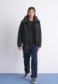 Ung mand går iført en sort jakke, sort skjorte, marineblå bukser og hvide sneakers foran en ensfarvet hvid baggrund.