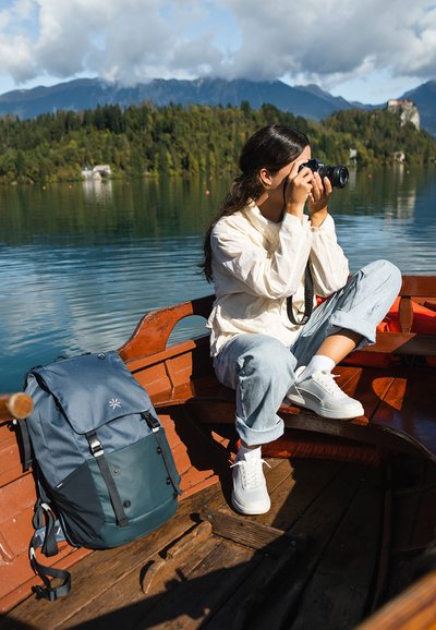 Mochila azul y gris sobre un bote de madera, con una persona sosteniendo una cámara, vestida con una chaqueta ligera y jeans. Escenario al aire libre de fondo.