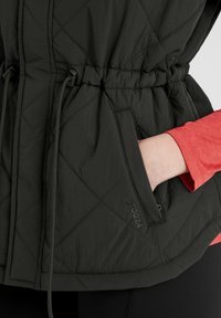 Black quilted jacket with cinched waist, vertical pocket, and logo detail. The fabric has a smooth texture, paired with a bright red shirt underneath.