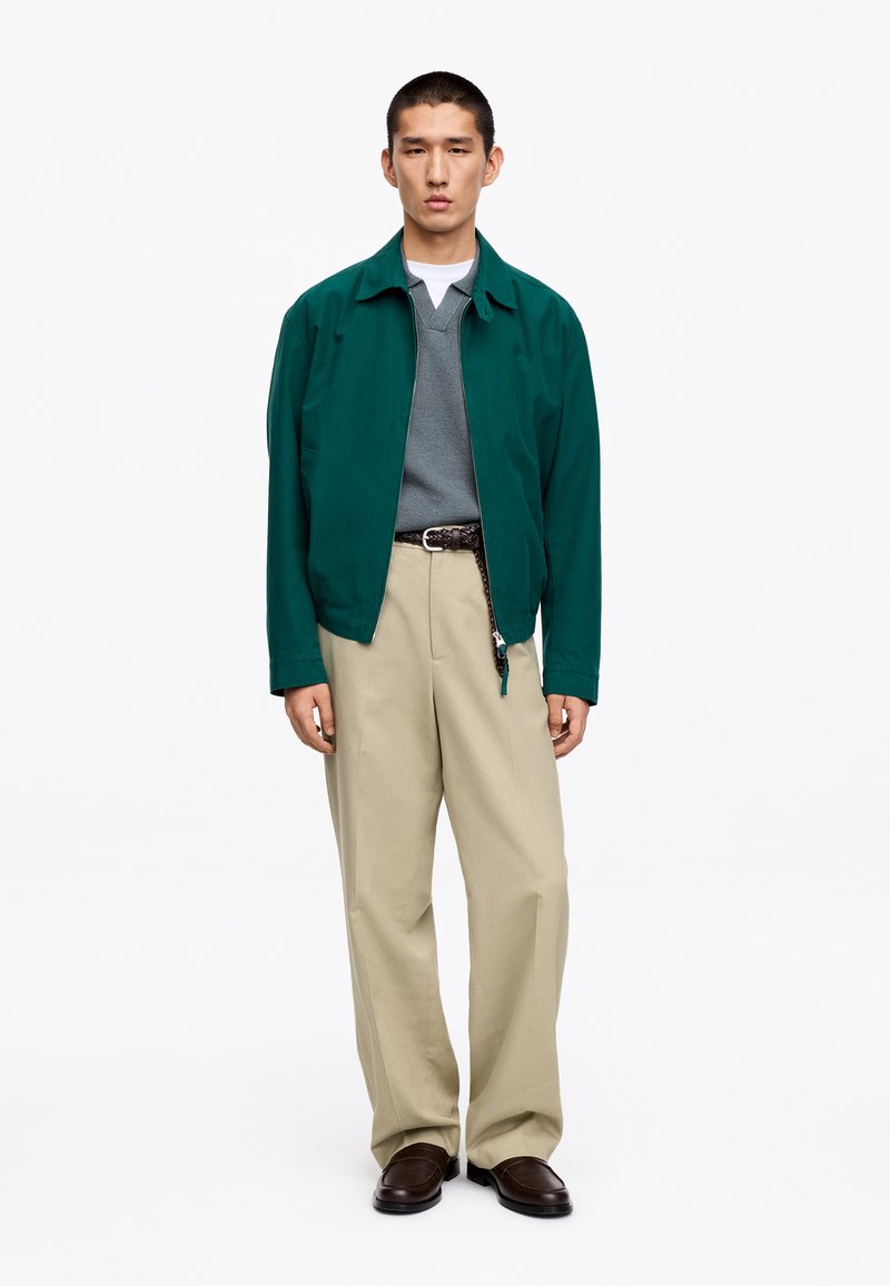 Jeune homme portant une veste verte, un pull gris col en V, un sous-maillot blanc, un pantalon beige, une ceinture tissée et des chaussures marron foncé, debout devant un fond blanc.