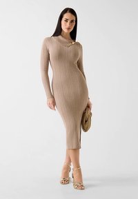 Robe midi en maille beige à texture côtelée verticale, avec un décolleté en V et un accent doré à l'épaule. Détail fente latérale visible.