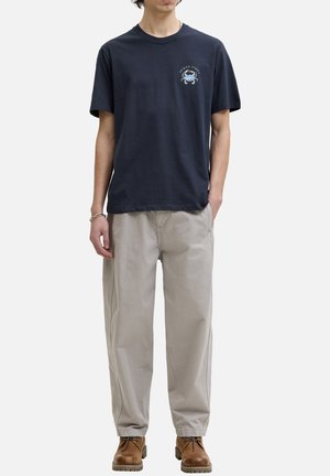 Persona con una camiseta azul marino con un cangrejo azul y el texto "Ocean Vibes", pantalones beige, botas marrones y una pulsera plateada, de pie con las manos en los bolsillos.