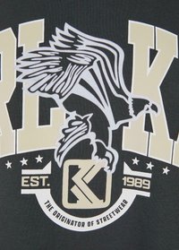 Sort stof med en stor grafik, der viser en ørn i hvid og guld. Teksten inkluderer "ETABL. 1989" og "OPRINDEREN AF STREETWEAR."