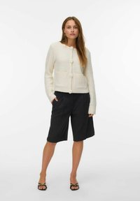 Kremfarget kort jakke med to lommer og runde knapper, sammen med mørke plisserte shorts og svarte sandaler med stropper.