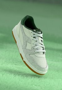 White sneakers with a green collar, suede-like texture, perforated detailing, and a rubber sole with orange accents. Classic lace-up design.