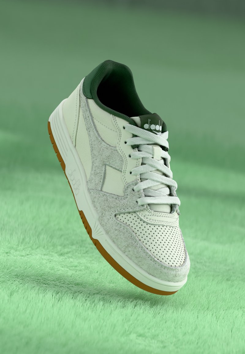 White sneakers with a green collar, suede-like texture, perforated detailing, and a rubber sole with orange accents. Classic lace-up design.