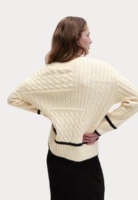 Maglione lavorato a maglia color crema con vari motivi a treccia, con polsini e orlo a costine delineati in nero. Vestibilità comoda.