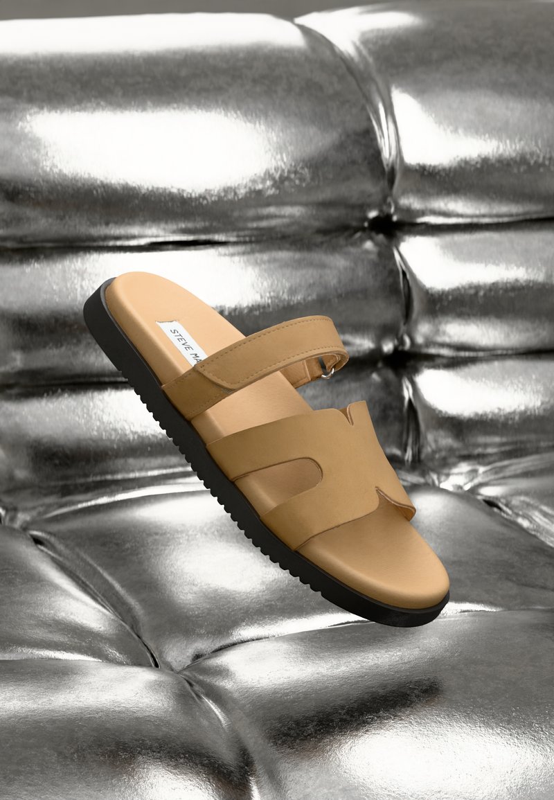 Tan slide sandals featuring two wide straps with cutouts, a smooth leather upper, and a textured black rubber sole, displayed against a metallic backdrop.