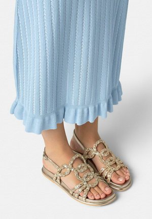 Voeten met gouden sandalen met schakels, gecombineerd met een lichtblauwe gebreide rok met geruffled zoom, op een lichtgrijze achtergrond.