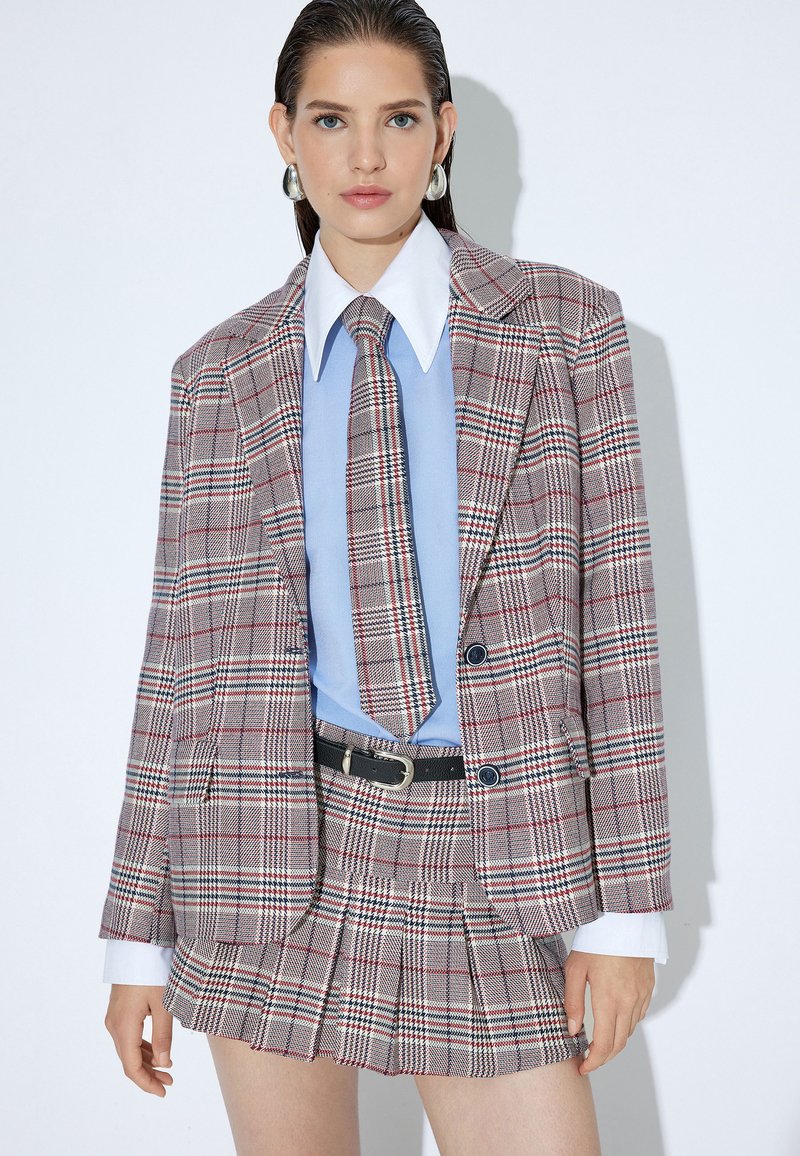 Geruite pak met een gestructureerde blazer met schoudervullingen, een geplooide mini-rok en een lichtblauw overhemd. Versterkt met een stropdas en een riem.