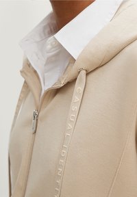 Beige rits-hoodie met een trekkoordkap, voorzien van de tekst "CASUAL IDENTITY" langs de trekkoorden en een witte overhemd eronder.