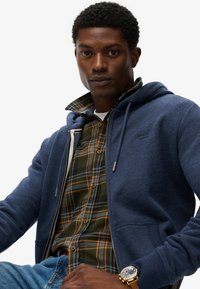 Giovane uomo che indossa una felpa blu con zip sopra una camicia a quadri verde, jeans blu e un orologio d'oro, seduto contro uno sfondo bianco semplice.