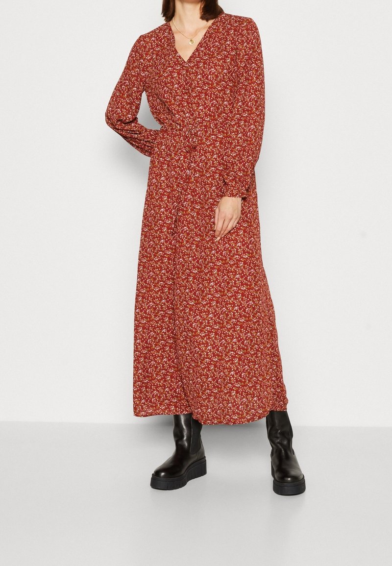 Femme portant une longue robe rouge fleurie à manches longues et des bottes à plateforme noires épaisses, debout devant un fond blanc uni.