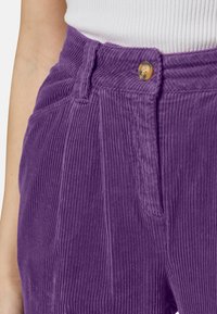 Pantaloni di velluto a coste viola con righe verticali, dotati di chiusura frontale con bottone e tasche laterali. Tessuto testurizzato e dettagli di taglio visibili.