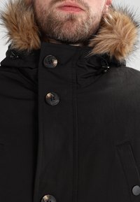 Svart parkajakke med høy krage, pelsforet hette og store svarte knapper. Har en teksturert overflate og sidelommer.