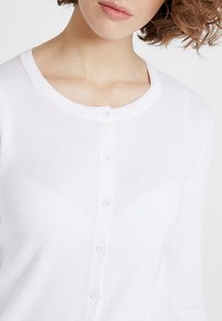 Bílý kardigan vyrobený z lehkého materiálu, s kulatým výstřihem, zapínáním na knoflíky a žebrovaným lemem. Hladká textura bez vzorů.