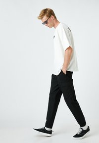 Witte oversized T-shirt met zwarte tekst, zwarte cargo pants en zwarte sneakers met witte zolen. Zonnebril en casual wandelhouding.