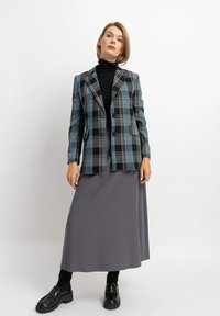 Karo-Blazer in Blau-, Schwarz- und Beigetönen, kombiniert mit einem langen grauen Rock. Der Blazer verfügt über einen Reverskragen und zwei Knöpfe. Schwarze Schuhe runden den Look ab.
