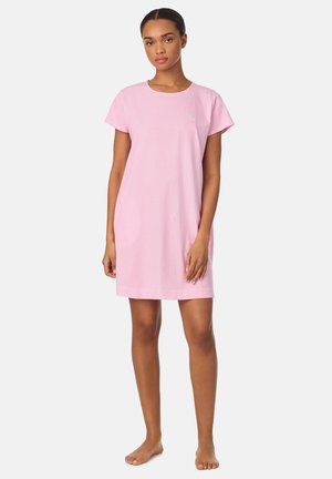 Vrouw staat blootsvoets en draagt een knielange jurk met korte mouwen in roze en witte strepen tegen een effen achtergrond.