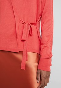 Cardigan en corail avec détail de taille nouée et poignet côtelé, associé à une jupe satin couleur rouille. Texture douce, design décontracté.