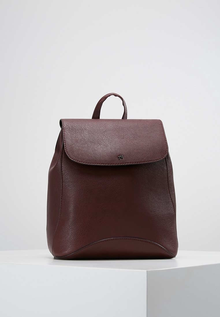 Zaino in pelle bordeaux con patta pieghevole, bordi arrotondati e manico superiore. Presenta una texture liscia e un logo in rilievo discreto sul davanti.