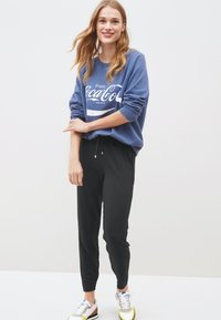 Blue sweatshirt with "Enjoy Coca-Cola" text, paired with black pants and multicolored sneakers. Casual fit with rolled sleeves and adjustable waistband.
