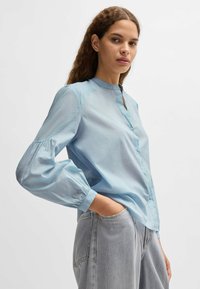 Jasnoniebieska bluzka z gładkiego materiału, z kołnierzykiem mandarynkowym, listwą guzikową i bufkowymi rękawami do trzech ćwierci. Noszona z szarymi dżinsami.