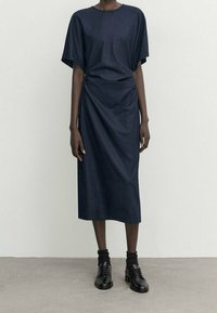 Navyblaues Kleid mit lockerer Passform, kurzen Ärmeln und asymmetrischem Drapé-Design. Mit glattem Stoff und knielangem Schnitt. Kombiniert mit schwarzen Schuhen.