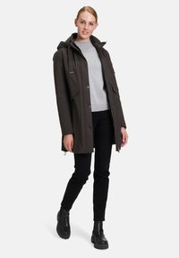 Parka imperméable marron foncé avec capuche, poches avant et fermeture éclair, portée par-dessus un pull gris clair et un pantalon noir.