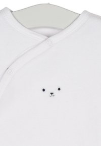 Une combinaison en coton blanc pour bébé avec des fermetures à pression, ornée d'un petit visage de ours noir brodé sur la poitrine. Texture douce, design simple.