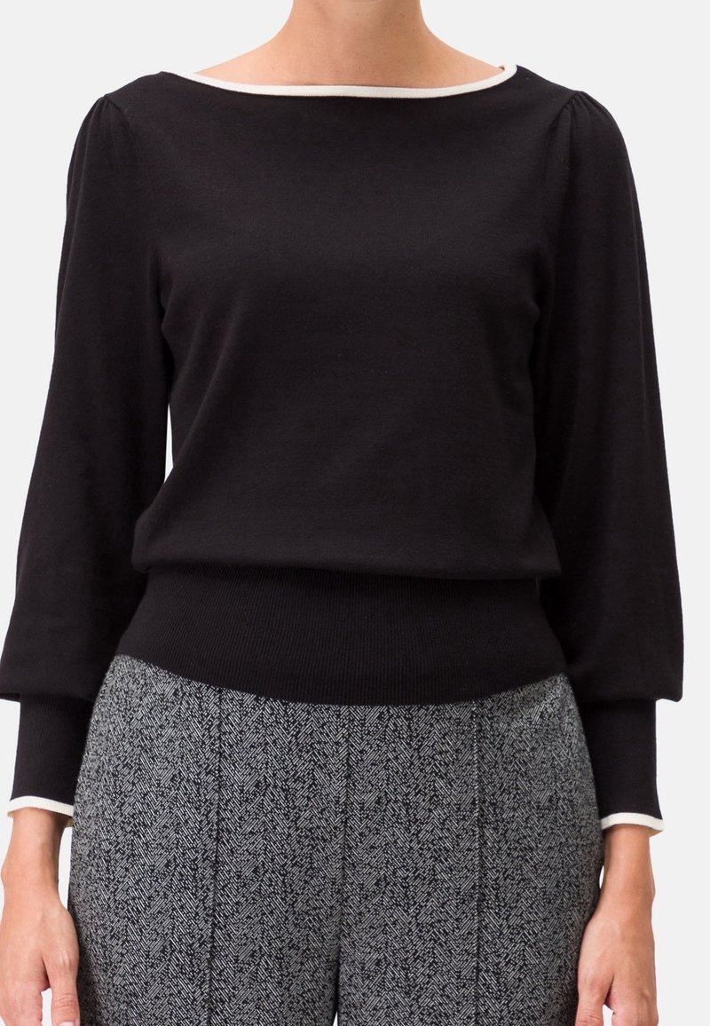 Femme portant un haut noir à manches longues avec un liseré blanc au col et aux poignets, associé à un pantalon gris à motifs taille haute.