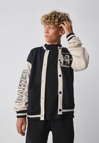 Chaqueta varsity negra y crema con mangas de color contrastante, cierre de botones y un parche con el logo en el pecho, con puños de canalé a rayas.