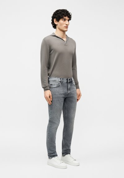 Άνδρας που στέκεται φορώντας πόλο με μακρύ μανίκι σε χρώμα του τάουπη, ξεθωριασμένο γκρι τζιν και άσπρα αθλητικά παπούτσια, μπροστά σε απλό ανοιχτό φόντο.