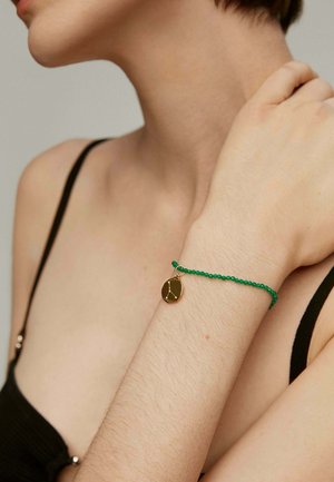 Pulsera de cuentas verdes con un charm circular dorado que presenta un diseño geométrico, llevada en la muñeca de una persona sobre un fondo neutral.