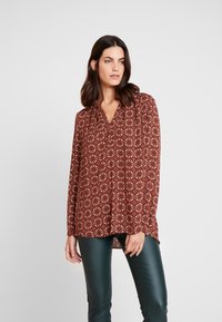 Blusa bordô com padrão, mangas longas, decote em V e corte solto; apresenta desenhos geométricos em creme e vermelho, combinada com calças verde-escuras.