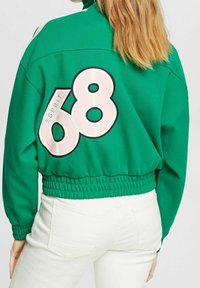 Grön kort sweatshirt med elastisk midja. Har en stor vit och rosa "68" grafik på baksidan. Tyget är mjukt med en livlig nyans.